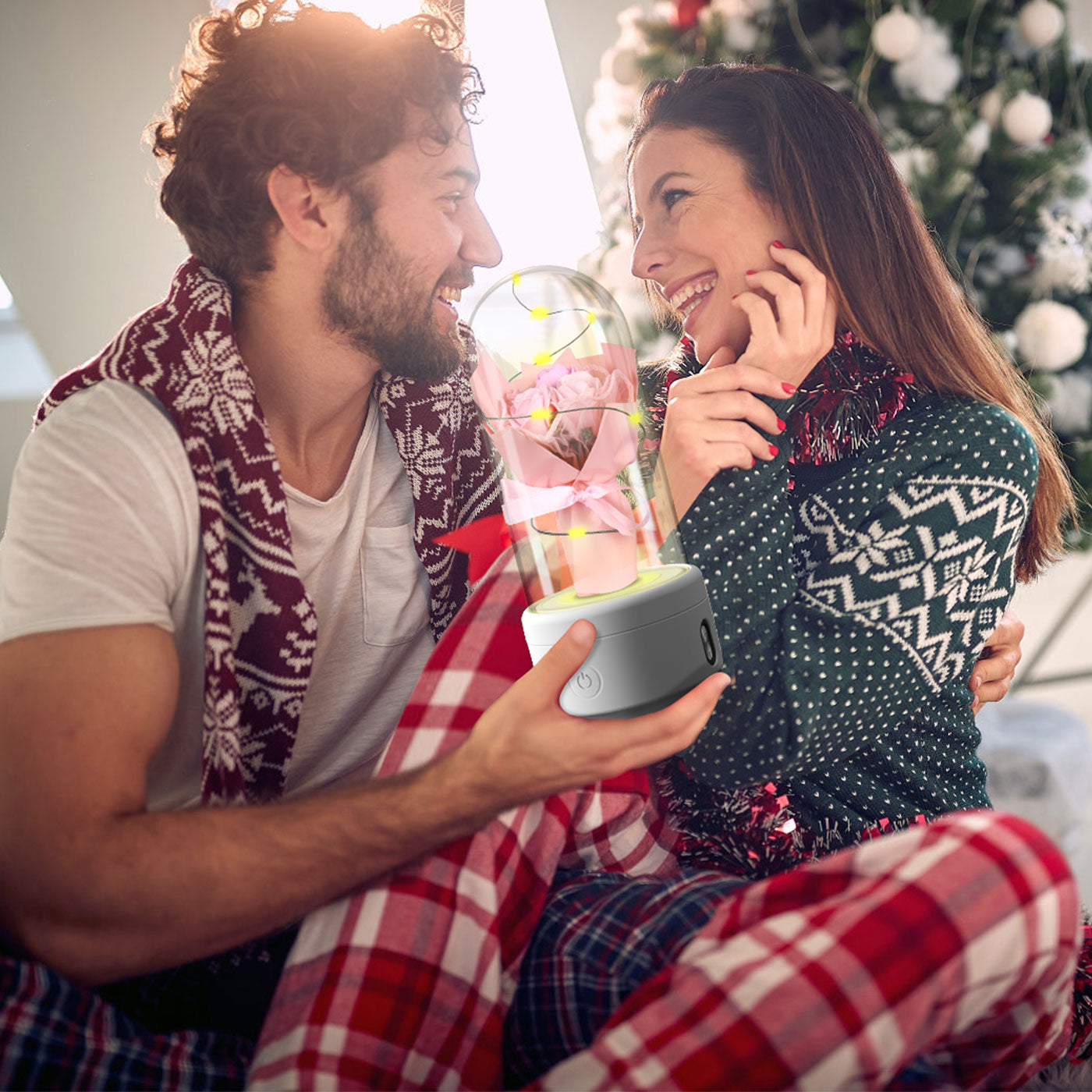 Couple smiling by Christmas tree holding rose lamp in glass dome, cozy holiday setting